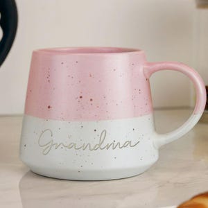 May include: A two-tone ceramic mug with a pink upper half and a white lower half. The word "Grandma" is written in cursive on the white section. The mug has a rounded handle and a speckled design.