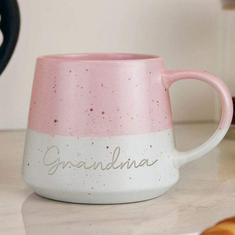 May include: A two-tone ceramic mug with a pink upper half and a white lower half. The word "Grandma" is written in cursive on the white section. The mug has a rounded handle and a speckled design.