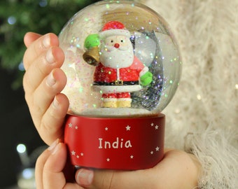 May include: A festive snow globe featuring Santa Claus in a red suit, holding a green bell. The globe sits atop a red base with the word "India" and star accents. The globe is filled with glitter.