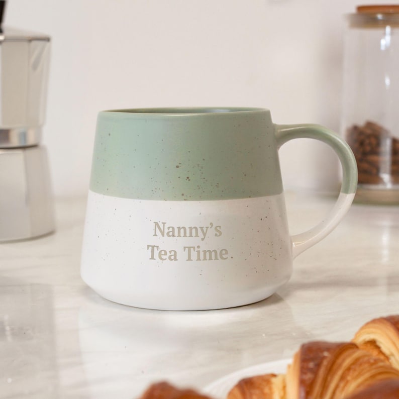 May include: A two-tone ceramic mug with a green top and white bottom. The mug has a curved handle and the words "Nanny's Tea Time" are printed on the front. The mug is on a white surface.