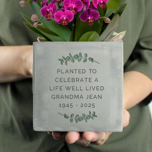 May include: A grey concrete planter holding a purple orchid. The planter has the inscription "PLANTED TO CELEBRATE A LIFE WELL LIVED GRANDMA JEAN 1945 - 2025" and is decorated with green leaf accents.