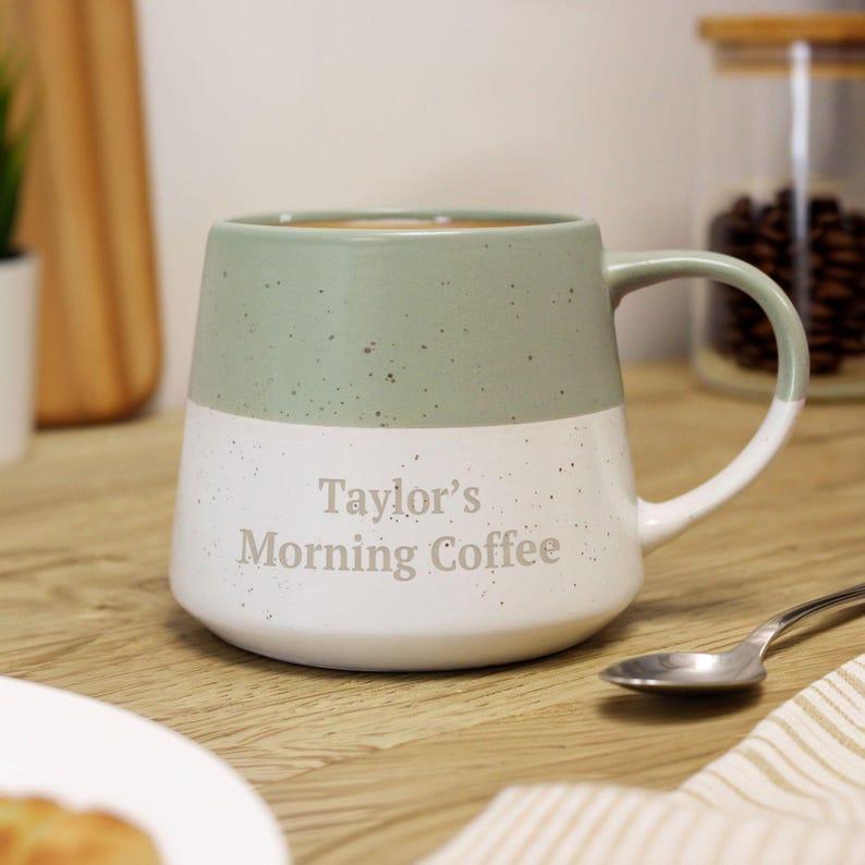 May include: A two-tone ceramic mug with a sage green top and white bottom. The mug has a speckled finish and a curved handle. The text "Taylor's Morning Coffee" is printed on the white section. A spoon and a napkin are next to the mug.
