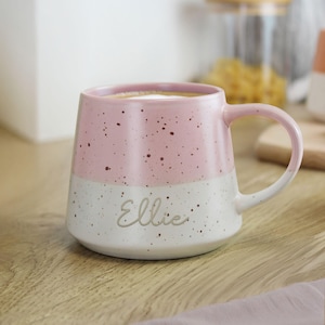 May include: A pink and white speckled ceramic mug with the name "Ellie" printed on the front. The mug is filled with a beverage and has a curved handle. The mug sits on a wooden surface.