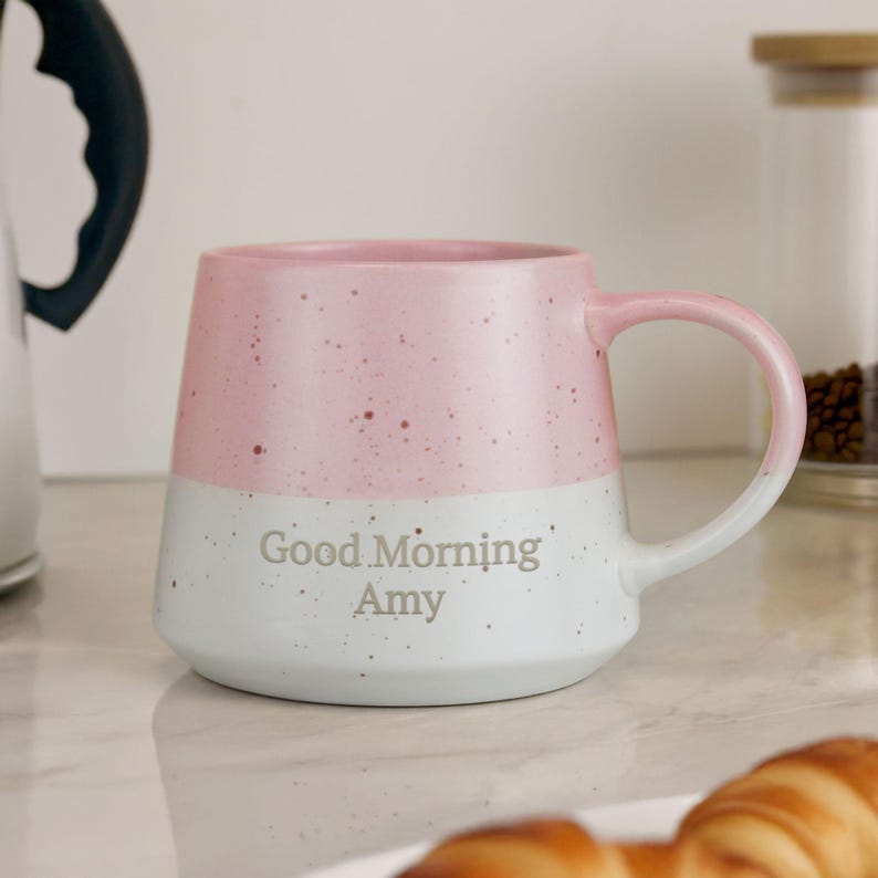 May include: A two-tone ceramic mug with a pink upper half and a white lower half, speckled with brown dots. The mug has a curved handle and the words "Good Morning Amy" are printed on the white section.