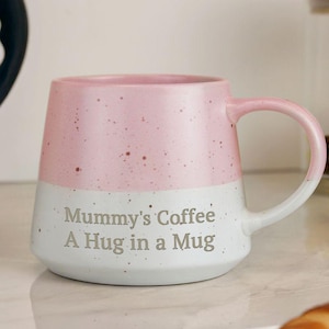 May include: A two-tone ceramic mug with a pink speckled top and a white speckled bottom. The mug has a curved handle and text that reads "Mummy's Coffee A Hug in a Mug".
