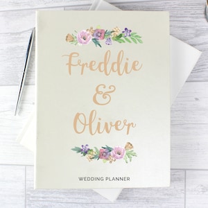 May include: A white wedding planner with a floral design and the names Freddie & Oliver in gold lettering. The text "Wedding Planner" is printed at the bottom.