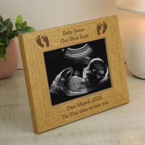 May include: A light oak picture frame displaying a sonogram image. The frame is engraved with the text "Baby Jones Our First Scan" and "Due March 2024 The First time we saw you". The frame has two baby footprint designs.