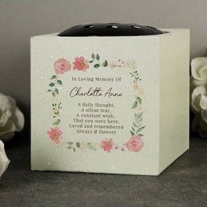 May include: A square, cream-coloured memorial vase with a black flower grid on top. The front is decorated with a floral wreath and the text "In Loving Memory Of Charlotte Anne" and a poem. The vase has a speckled finish.
