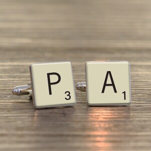 May include: A pair of silver cufflinks with white Scrabble tiles. The tiles spell "PA" with the letter P having a score of 3 and the letter A having a score of 1.