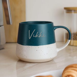 May include: A teal and white ceramic mug with the name "Vikki" in a script font. The top half of the mug is teal, and the bottom half is white with speckled detailing. The handle is white with a teal accent.