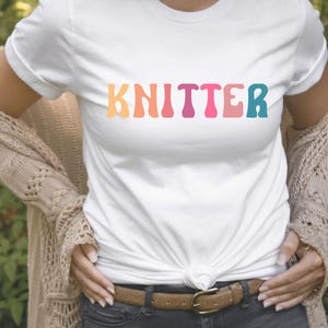 Puede incluir: Una camiseta blanca con la palabra "KNITTER" impresa en colores arco&iacute;ris. La camiseta la lleva puesta una persona con un c&aacute;rdigan marr&oacute;n y unos vaqueros azules.