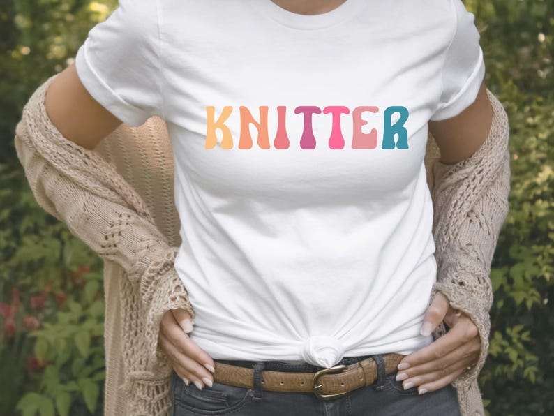 Puede incluir: Una camiseta blanca con la palabra "KNITTER" impresa en colores arco&iacute;ris. La camiseta la lleva puesta una persona con un c&aacute;rdigan marr&oacute;n y unos vaqueros azules.