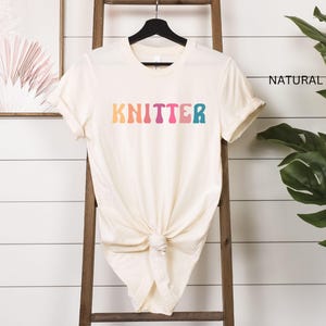 Puede incluir: Una camiseta blanca con la palabra "KNITTER" en colores arco&iacute;ris. La camiseta est&aacute; colgada en una escalera de madera.