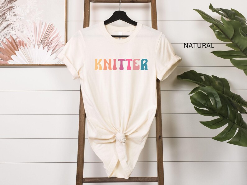 Puede incluir: Una camiseta blanca con la palabra "KNITTER" en colores arco&iacute;ris. La camiseta est&aacute; colgada en una escalera de madera.