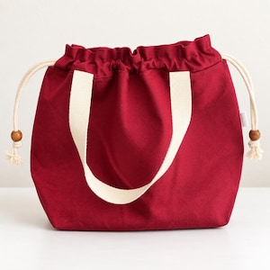 May include: A deep red drawstring bag featuring two cream-coloured fabric handles and a matching cream drawstring cord. The cord is threaded through the top opening and finished with small, round, brown wooden beads at each knotted end.