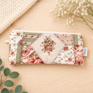 May include: A rectangular zipped pouch featuring a patchwork design of floral and striped patterns in shades of red, green, and cream. The centre panel displays a diamond shape with a floral bouquet motif. A small white tag reads The Wool N Frog.