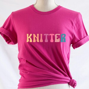 Puede incluir: Una camiseta rosa con la palabra "KNITTER" impresa en colores arco&iacute;ris.