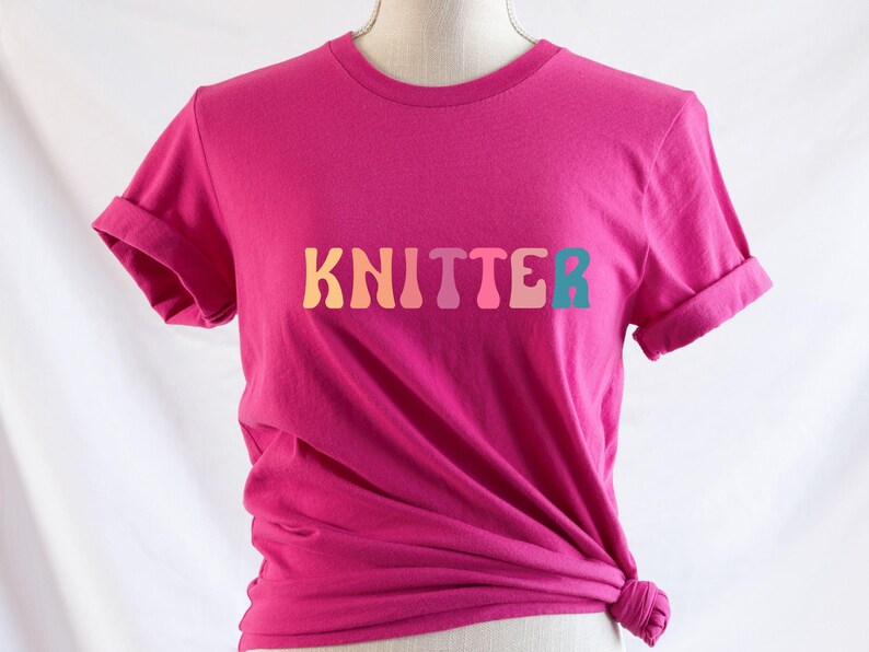 Puede incluir: Una camiseta rosa con la palabra "KNITTER" impresa en colores arco&iacute;ris.