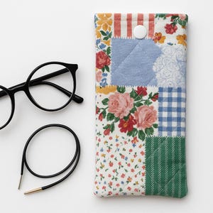 May include: A multi-patterned glasses case with a button fastening, showcasing floral, striped, and gingham designs in red, blue, green, and white. Black round-frame spectacles and a black eyeglass cord are next to the case.