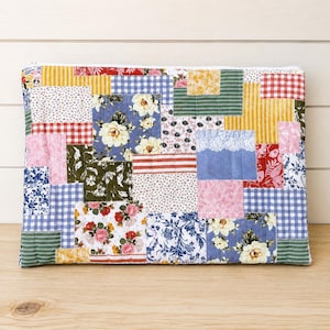May include: A rectangular quilted pouch featuring a patchwork design of various fabric squares. The patterns include blue and white gingham, yellow stripes, red and white checks, and floral prints in shades of pink, blue, green, and yellow.