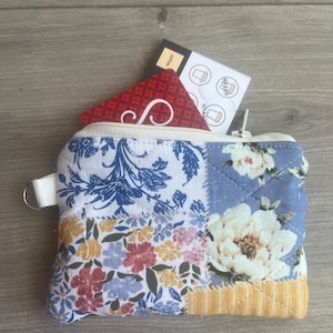 May include: A small, quilted coin purse with a floral patchwork design. The purse features a zipper closure, a white loop, and a red card with a white design peeking out. The interior holds a small card and a rewards card.
