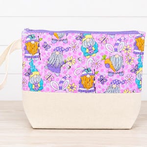 Könnte beinhalten: Eine Reißverschlusstasche aus rosa und violett gepunktetem Stoff mit einem sich wiederholenden Muster aus Gnomen, Schmetterlingen und Blumen. Der obere Teil ist gemustert, während der untere Teil aus einfarbigem, cremefarbenem Canvas besteht.