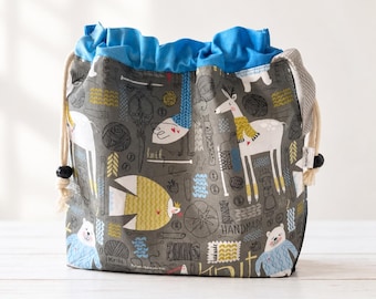 May include: A drawstring project bag featuring a gray background with a repeating pattern of deer, bears, scissors, yarn balls, and the word "knit". The bag has a bright blue fabric top section and thick cream-colored rope drawstrings on each side.