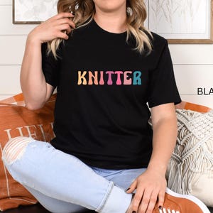 Puede incluir: Una camiseta negra con la palabra "KNITTER" en colores arco&iacute;ris. La camiseta la lleva puesta una persona sentada en un sof&aacute;.