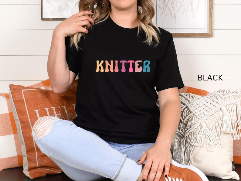 Puede incluir: Una camiseta negra con la palabra "KNITTER" en colores arco&iacute;ris. La camiseta la lleva puesta una persona sentada en un sof&aacute;.