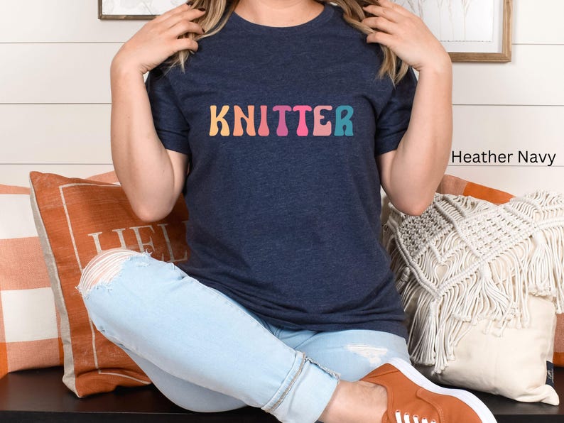 Puede incluir: Una mujer lleva una camiseta azul marino jaspeada con la palabra "KNITTER" impresa en colores arco&iacute;ris. Est&aacute; sentada en un sof&aacute; con cojines y lleva unos vaqueros azules claros.