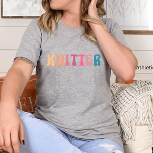 Puede incluir: Una camiseta gris con la palabra "Knitter" impresa en colores arco&iacute;ris. La camiseta est&aacute; hecha de un material suave y c&oacute;modo y es perfecta para el uso diario.