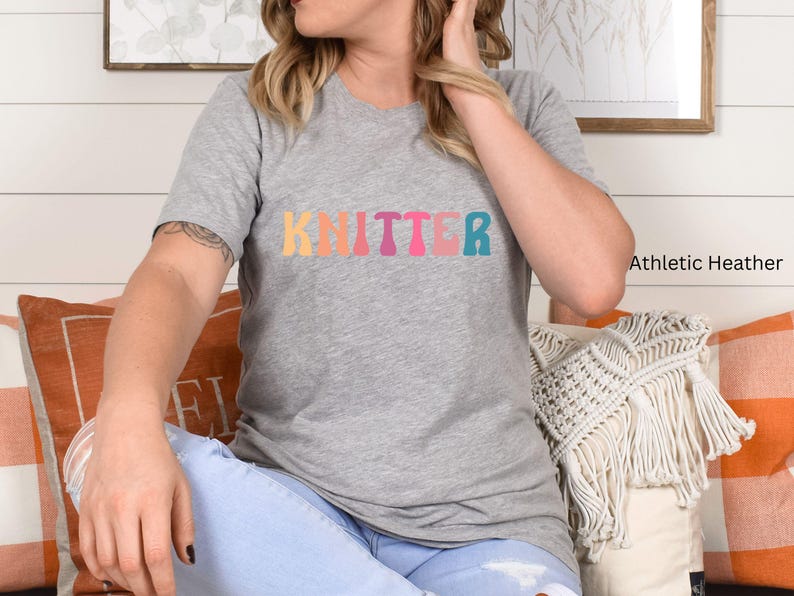 Puede incluir: Una camiseta gris con la palabra "Knitter" impresa en colores arco&iacute;ris. La camiseta est&aacute; hecha de un material suave y c&oacute;modo y es perfecta para el uso diario.