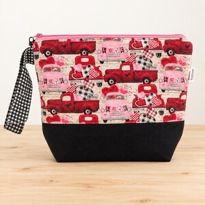 May include: A zippered pouch featuring a pattern of red and pink vintage trucks carrying hearts, with a black fabric base and a black and white gingham wrist strap. The trucks display the word LOVE on their front bumpers amidst heart motifs.