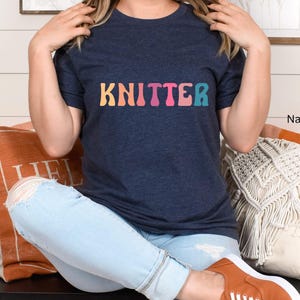 Puede incluir: Una mujer lleva una camiseta azul marino con la palabra "KNITTER" en colores arco&iacute;ris. Est&aacute; sentada en un sof&aacute; con almohadas y lleva unos vaqueros azules claros.