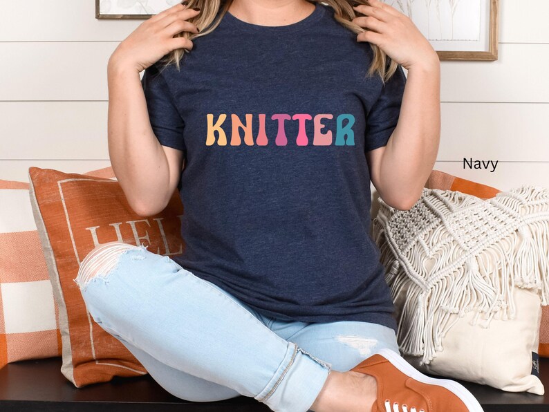 Puede incluir: Una mujer lleva una camiseta azul marino con la palabra "KNITTER" en colores arco&iacute;ris. Est&aacute; sentada en un sof&aacute; con almohadas y lleva unos vaqueros azules claros.