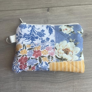 May include: A small, quilted coin purse with a zip closure and a white wrist strap. The patchwork design features floral patterns in blue, yellow, pink, and white. The purse is approximately 13 cm wide and 10 cm tall.