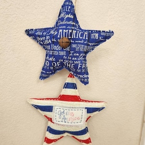 May include: Two star-shaped patriotic ornaments. The top star is blue with white text and a small brown ball. The bottom star has red and blue stripes with the text "God Bless America". Both are hanging by twine.
