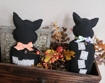 May include: Two black cat-shaped plush dolls with pointed ears and bow ties, displayed in a rustic wooden drawer. One cat has an orange and white bow tie, the other a green and purple bow tie. The dolls have stitched letters on their bodies.