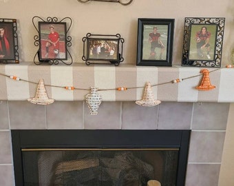 May include: A Halloween-themed mantel decoration featuring a garland with orange witch hats and white hats, strung with beads. The mantel is decorated with framed photos and a decorative vase. The color scheme includes orange, white, and neutral tones.
