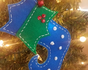 May include: A blue felt elf shoe ornament featuring white stitched edges and small white dots. The top is decorated with a green felt holly leaf shape, three small red berries, and a single red jingle bell attached to the upper corner.