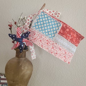 May include: A decorative patriotic arrangement featuring a fabric flag with red, white, and blue patterns, lace trim, and a wooden stick. The flag is displayed in a brown vase with a pinwheel and floral accents, perfect for Independence Day.