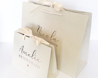 May include: Two beige gift bags with cream ribbon handles. The larger bag has the name "Nicola" printed in gold script. The smaller bag has the name "Amelia" and the word "Bridesmaid" printed in rose gold script, with a small heart.