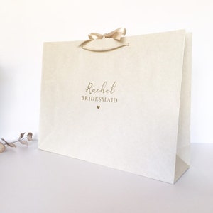 May include: Cream gift bag with a satin ribbon bow. The bag features the name "Rachel" and the word "Bridesmaid" in gold script, with a small heart beneath. The bag is on a white surface.