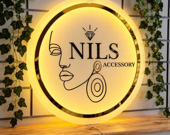 May include: A circular, illuminated wall sign featuring a black line-art profile of a face wearing a large earring. Above the face is a diamond icon, and the text reads NILS ACCESSORY in black lettering. The sign has a gold-toned metallic border.