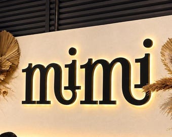 May include: A wall-mounted sign displaying the word mimi in bold black lowercase letters with a warm backlit glow. The sign is flanked by two large arrangements of dried palm leaves, pampas grass, and seed pods, under two beige fabric pendant lights.