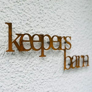 May include: A rusty brown metal sign that reads "keepers barn" mounted on a textured white wall. The sign is in a stylized font, with the letters connected. The sign is attached to the wall with small nails.