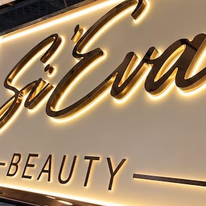 May include: A rectangular wall sign featuring the gold-colored script text Si'Eva above the word BEAUTY in a sans-serif font. The sign has a white background and is illuminated from behind the raised letters, creating a warm glowing halo effect.