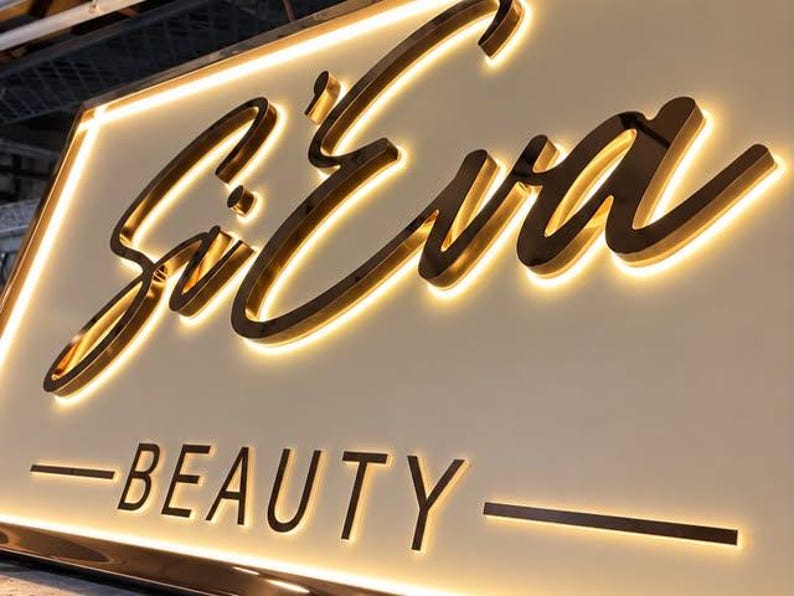 May include: A rectangular wall sign featuring the gold-colored script text Si'Eva above the word BEAUTY in a sans-serif font. The sign has a white background and is illuminated from behind the raised letters, creating a warm glowing halo effect.
