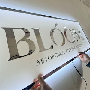 Puede incluir: Un letrero rectangular con la palabra "BLOOM" en letras plateadas reflectantes grandes. Debajo, un texto más pequeño dice "АВТОРСЬКА СТУДІЯ КВІТІВ". El letrero es blanco con un borde plateado y está siendo sostenido por dos manos.
