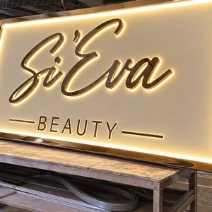 May include: A rectangular, wall-mounted sign with a gold-toned metallic frame and a white face. The sign features the text Si'Eva in a large, dark, cursive script and the word BEAUTY in a smaller, sans-serif font, both illuminated by backlighting.